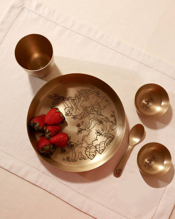 A brass thali set with etched jungle-themed patterns, including a plate, two bowls, a spoon, and a glass, displayed on a white surface with strawberries placed on the plate.
